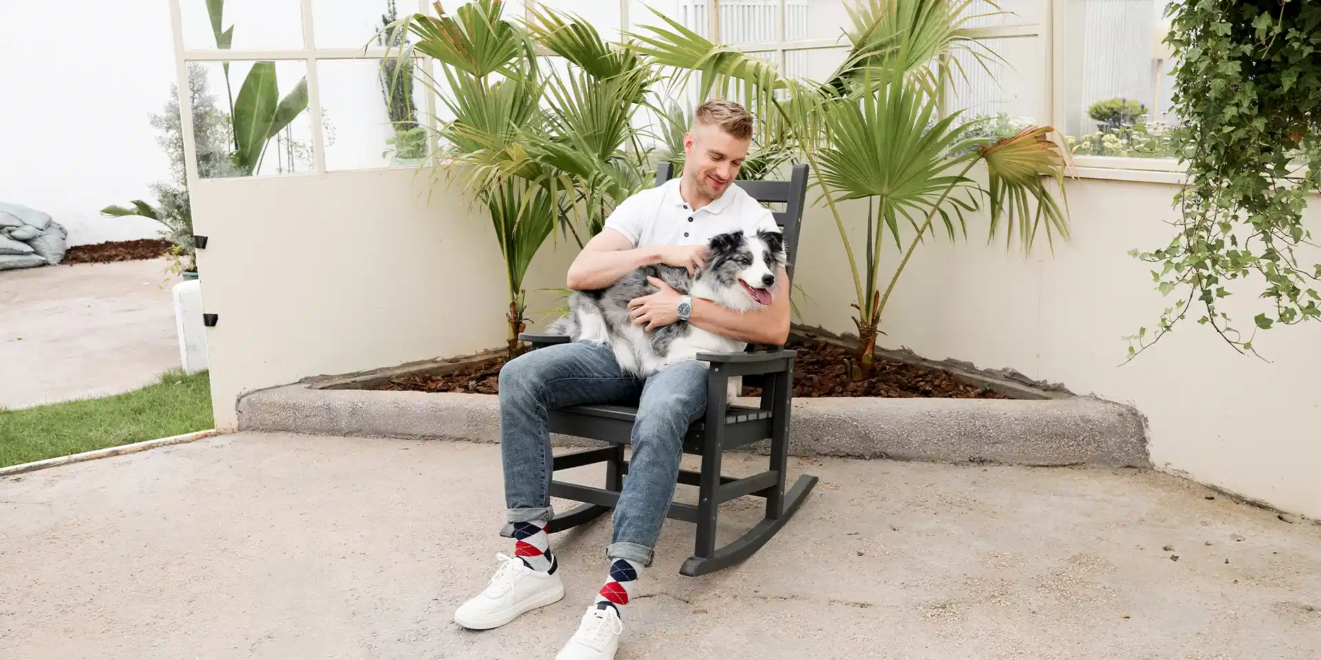 A man sits on the front porch rocking chair with his dog